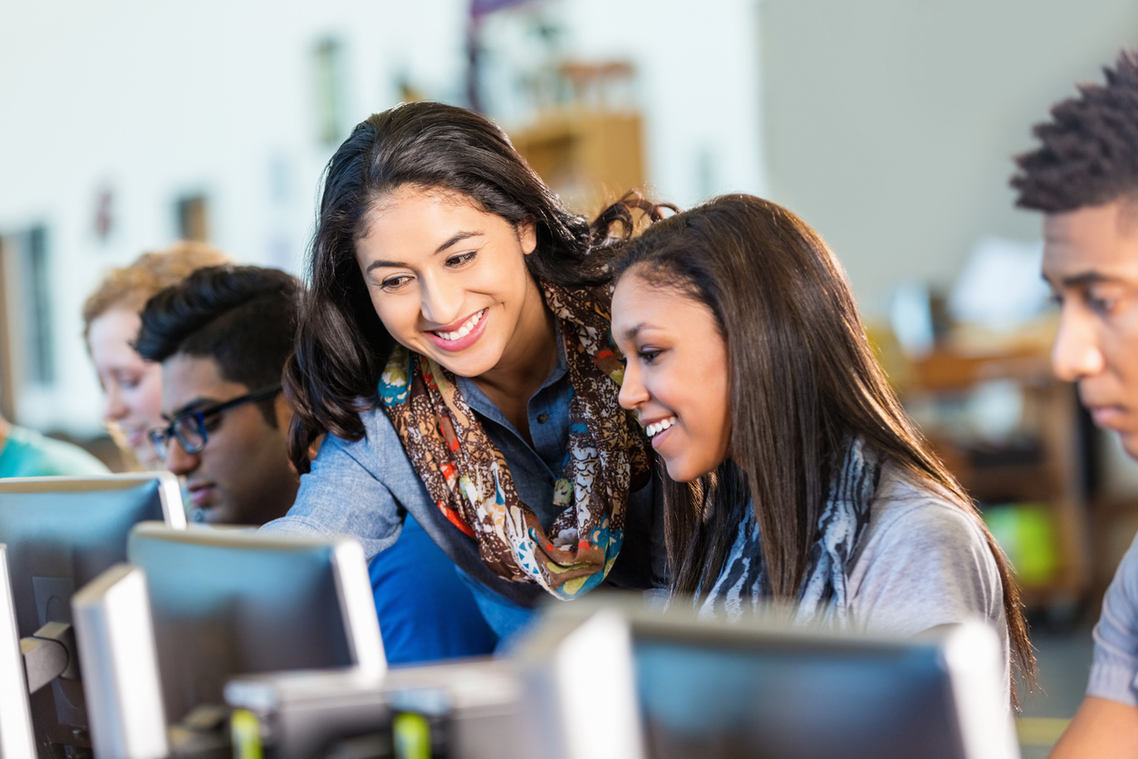 Happy Hispanic teacher assisting high school students with computers.